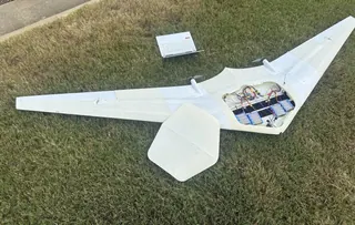 White 3D-printed flying wing with two propellers on grass, hatch removed to show wiring and blue battery packs; a laptop sits nearby