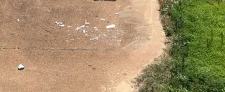 Scattered white 3D-printed fragments on weathered pavement beside a grassy verge, seen from above