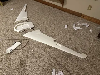 Disassembled white 3D-printed flying wing on carpet: two wing panels, a center section with an oval bay, and a control surface with a servo