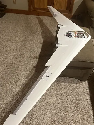 Assembled white swept flying wing on carpet with the center hatch open, showing a battery pack, wiring and two rear motors