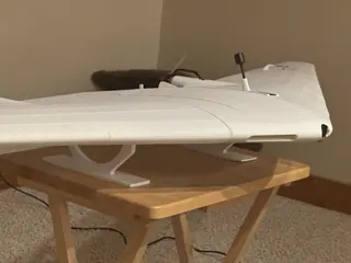 Side view of a white 3D-printed flying wing on white stands atop a wooden folding table, with a propeller and short black antenna