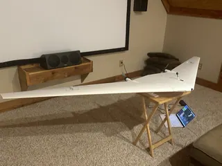 White swept flying wing with twin rear motors resting on a folding wooden tray table in a carpeted room, a laptop on the floor