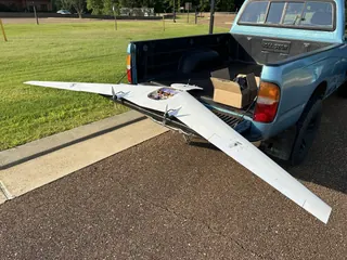 White swept flying wing resting across the tailgate of a light-blue pickup, wingtips extending well past the truck, hatch open