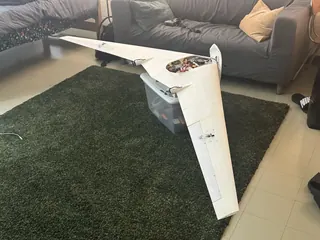 White flying wing propped on a clear storage bin on a dark green rug, hatch open with wiring visible