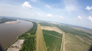 Onboard wide-angle view from an aircraft banking over farm fields, a levee road and a wide brown river with sandbars