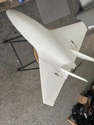 High-angle view of a white 3D-printed jet-style delta with twin canted fins and a rear propeller, on a wooden stool