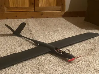 Dark grey 3D-printed motor glider with a long rounded-tip wing, slim tail boom and red nose propeller, open hatch showing wires, on carpet