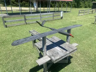 Dark grey motor glider with a long rounded-tip wing, slim tail boom and red propeller on a wooden stand at a grass flying field