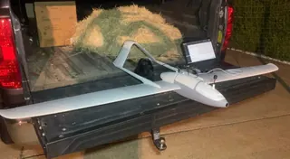 White twin-boom UAV with an antenna mast on a pickup tailgate at night, beside a laptop showing a map and FPV goggles